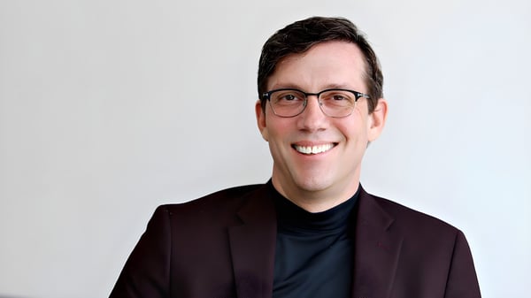 Portrait of a man wearing glasses, a black turtleneck, and a dark blazer against a plain white background.