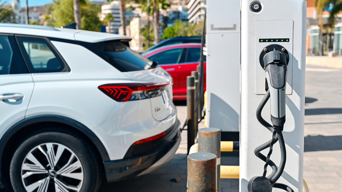 A white electric SUV parked next to a public charging station on a sunny city street with palm trees in the background