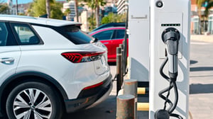 A white electric SUV parked next to a public charging station on a sunny city street with palm trees in the background