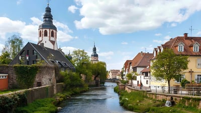 Historic German town with a river, stone walls, and traditional architecture featuring church steeples under a blue sky.