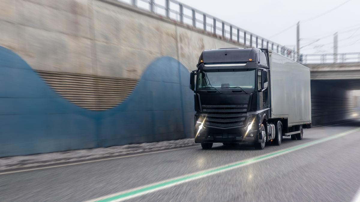 A black semi-truck with a white trailer driving on a highway next to a concrete wall and underpass