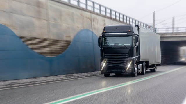 A black semi-truck with a white trailer driving on a highway next to a concrete wall and underpass