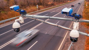 Overhead gantry with multiple surveillance cameras and speed detection units monitoring a multi-lane highway with blurred cars.