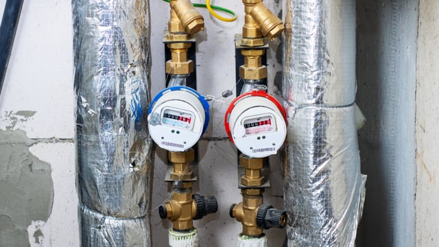 Two water meters with blue and red rings mounted on brass pipework between foil-wrapped insulated pipes on a utility room wall