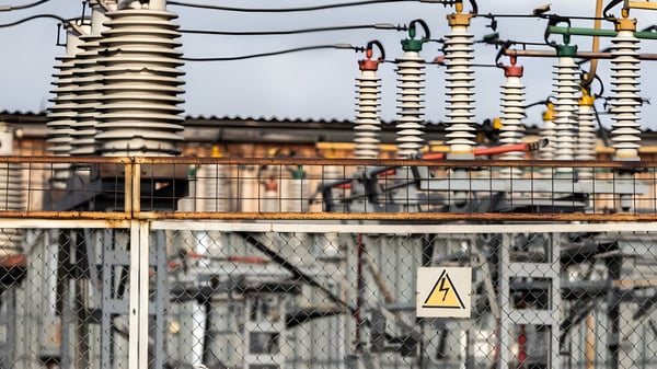 High-voltage ceramic insulators and power lines at an electrical substation behind a chain-link fence with a warning sign.