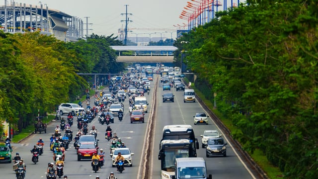 Busy multi-lane highway in a city with cars, trucks, and many motorbikes, flanked by green trees and buildings.