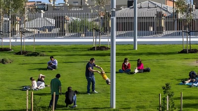 People relaxing on a green lawn in an urban park with smart lighting poles and a metal fence in the background.