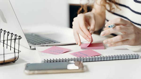 Close-up of a person's hands placing pink adhesive notes on a spiral notebook next to a laptop and smartphone.