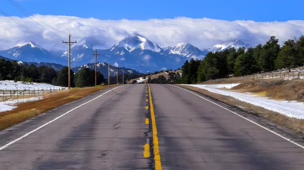 Two-lane asphalt highway with utility poles leading toward snow-capped mountains under a blue sky.