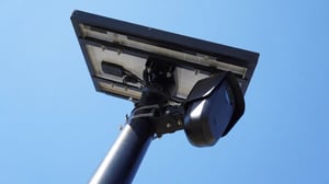Low-angle shot of a solar-powered Flock Safety license plate reader camera mounted on a utility pole against a blue sky