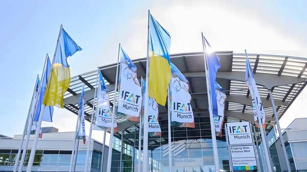 IFAT Munich and Ukrainian flags flying in front of the Messe München exhibition center entrance under a clear sky.
