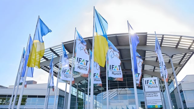 IFAT Munich and Ukrainian flags flying in front of the Messe München exhibition center entrance under a clear sky.