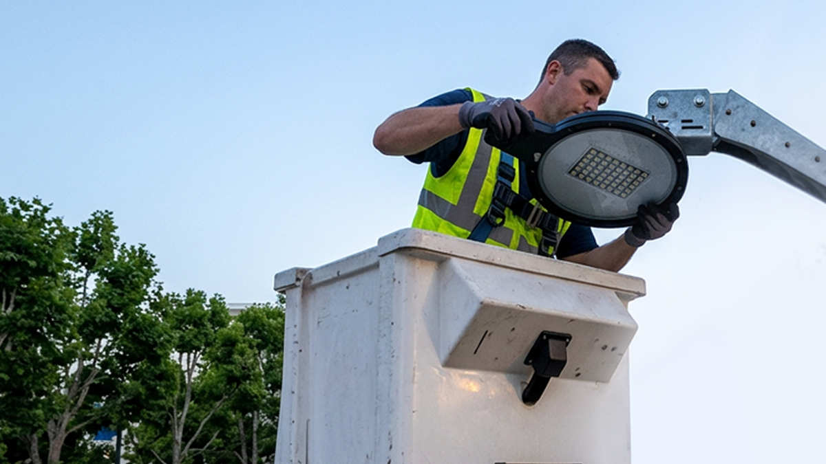 A technician in a cherry picker bucket installs a circular LED streetlight fixture against a clear sky