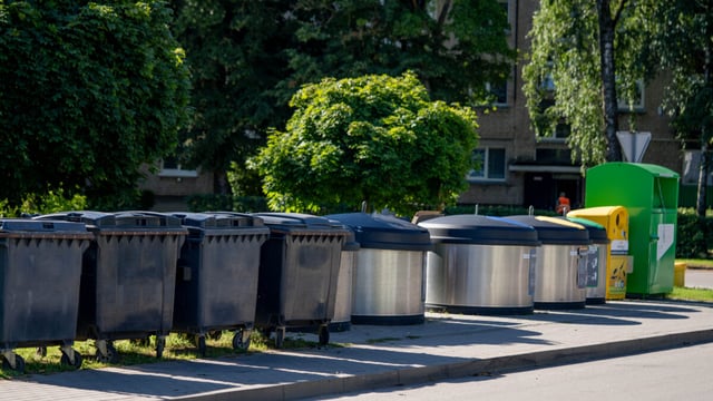 Row of dark wheeled waste containers and steel semi-underground bins on a paved pavement in front of trees and apartment blocks