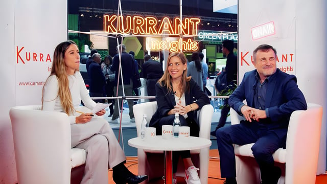 Three people sit in white armchairs for a Kurrant Insights interview at a busy indoor trade show.
