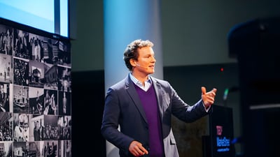 John Paul Farmer speaking on a stage next to a collage of black and white photographs.