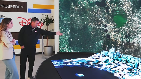 Two people view a 3D digital twin model of Orlando on a large display table in front of a Disney branded wall.