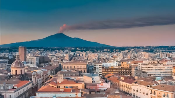 Aerial view of Catania, Italy, with Mount Etna emitting a plume of smoke in the background at dusk.