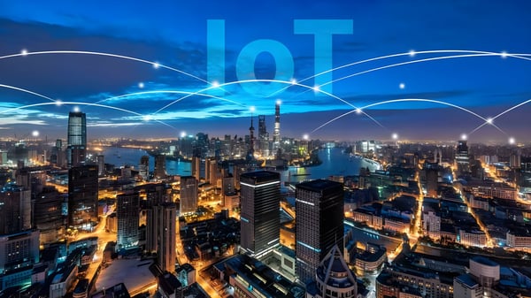 Aerial view of a lit city skyline at night with digital network lines and IoT text overlaying the sky.