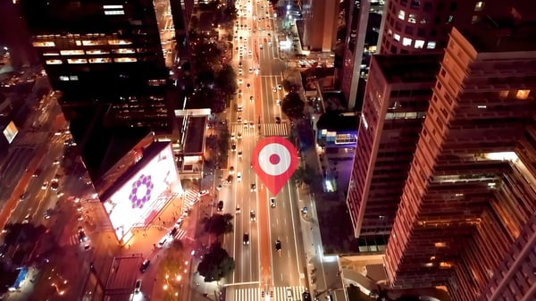 Aerial view of a city street at night with illuminated buildings, traffic, and a digital red location pin overlay.