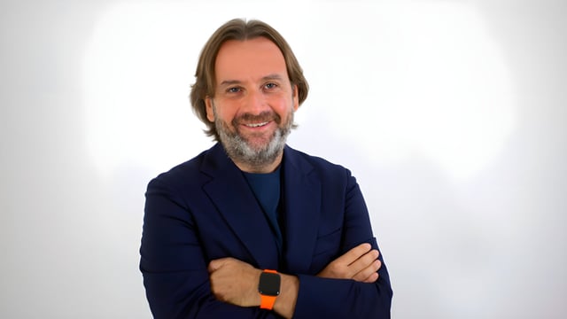 Portrait of Gianluca Longo wearing a navy blazer and an orange smartwatch against a white background.