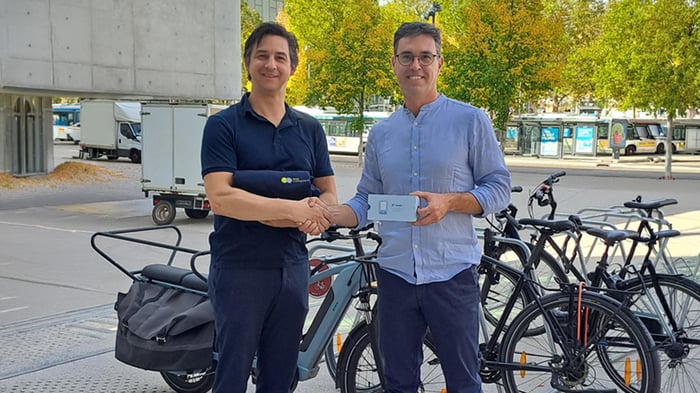 Two men shaking hands outdoors, one holding a small white box, with a cargo e-bike and several bicycles parked nearby.