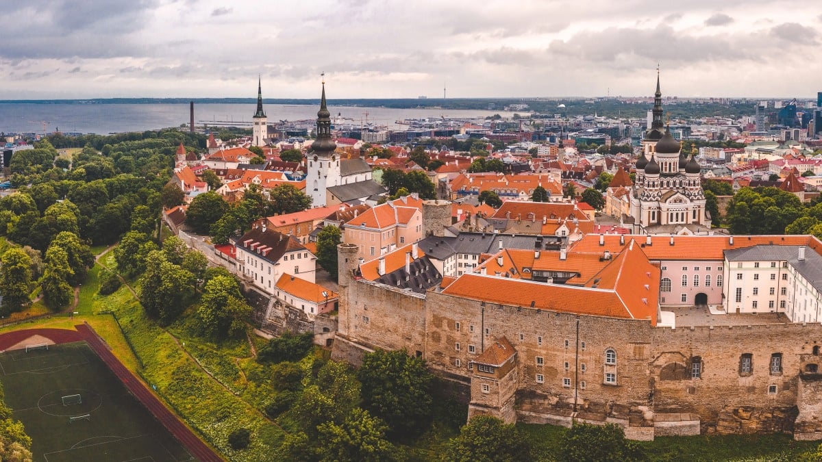 Aerial panoramic view of Tallinn's Old Town with historic buildings, spires, red roofs, a stadium, and a harbor in the distance.