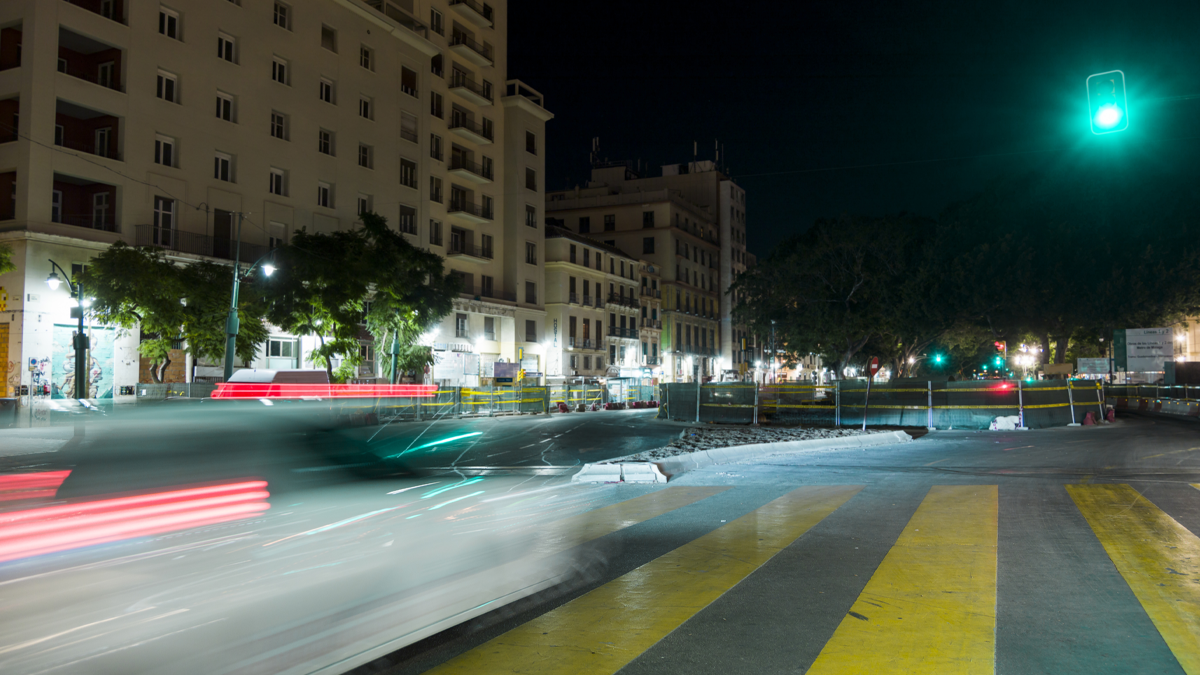Long exposure night photograph of a city street with blurred car light trails, a yellow crosswalk, and buildings.