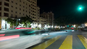 Long exposure night photograph of a city street with blurred car light trails, a yellow crosswalk, and buildings.