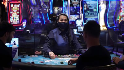 A masked dealer stands behind a plexiglass barrier at a Las Vegas casino table with players and slot machines.