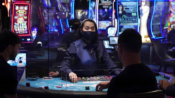 A masked dealer stands behind a plexiglass barrier at a Las Vegas casino table with players and slot machines.