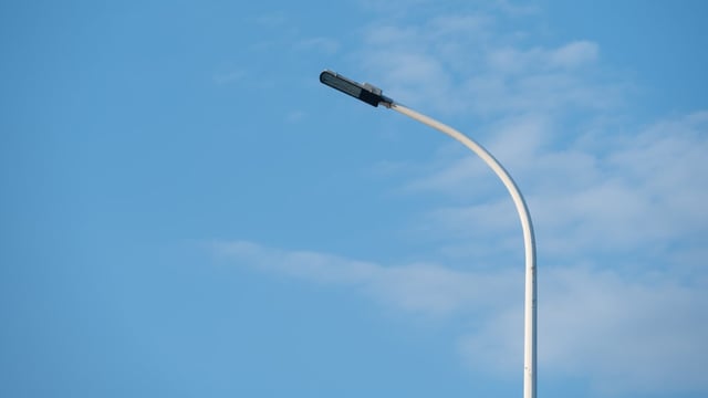 A white curved-arm street lamp with an LED luminaire head set against a clear blue sky with thin clouds