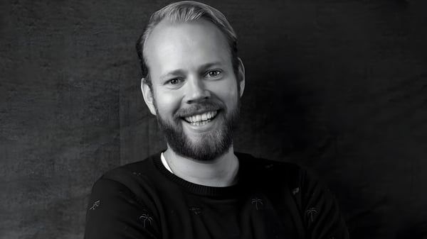 Black and white headshot of a smiling man with a beard wearing a dark sweater against a textured background.