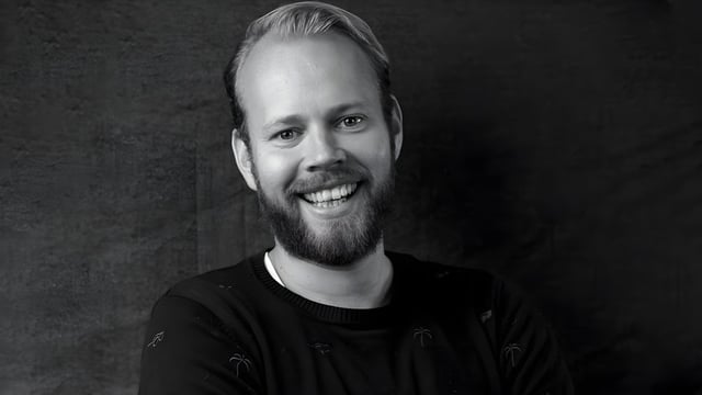 Black and white headshot of a smiling man with a beard wearing a dark sweater against a textured background.