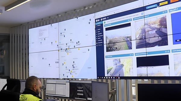 Operator in a high-visibility vest monitors a large video wall displaying city maps and live surveillance camera feeds.