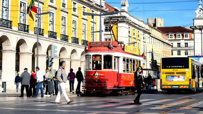 Pedestrians cross a street in Lisbon near a red vintage tram and a yellow bus by the Praça do Comércio.