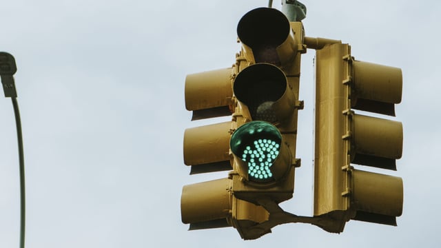 Yellow overhead traffic signal showing a green light on a mast arm beside a street lamp against an overcast sky