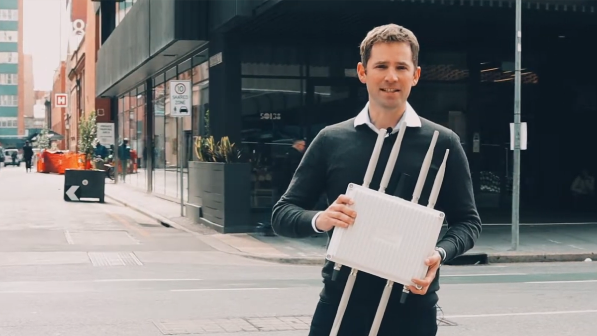 A man stands on a city street holding a white V2X communication device with multiple antennas.
