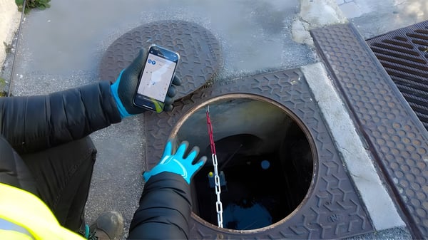 A worker in blue gloves holds a smartphone over an open manhole with a sensor suspended by a chain inside.