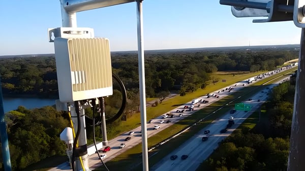 Wireless gateway mounted on a high pole overlooking a multi-lane highway with traffic and surrounding forest.