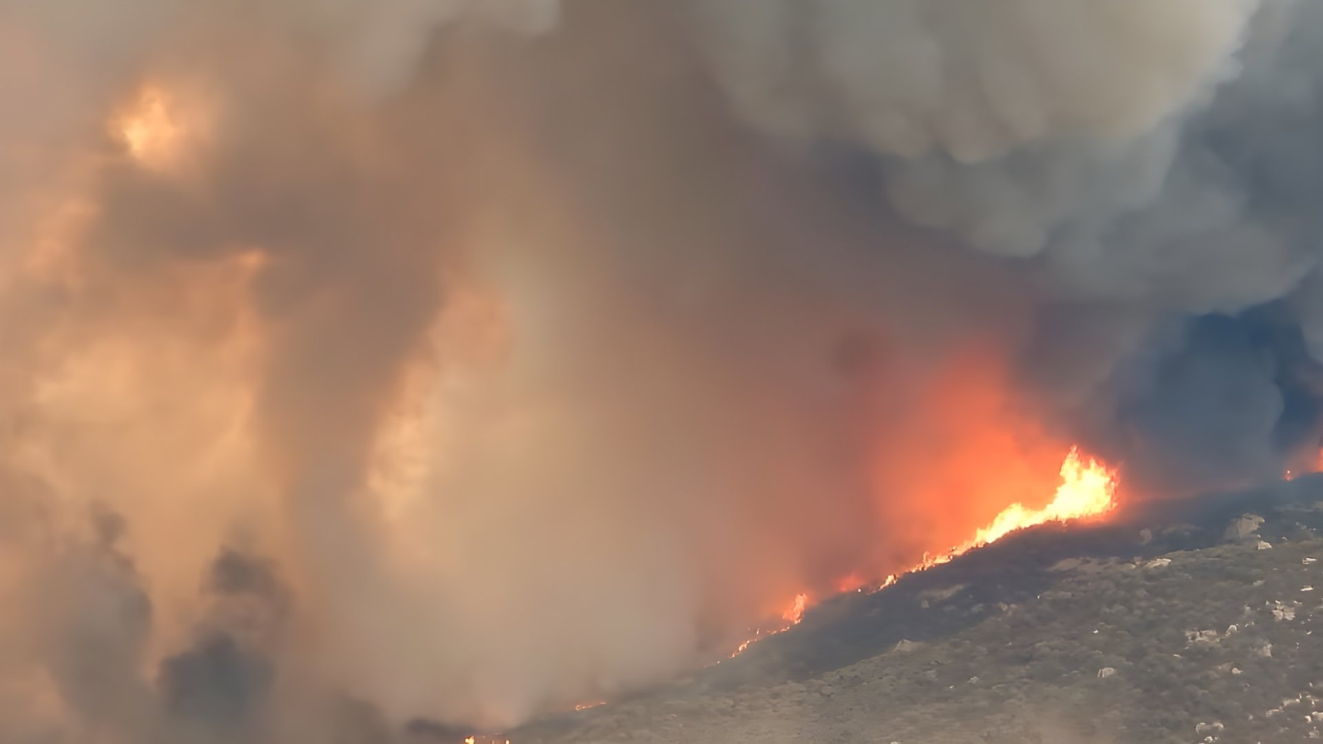 Thick plumes of orange and grey smoke rising from a wildfire burning across a hillside.