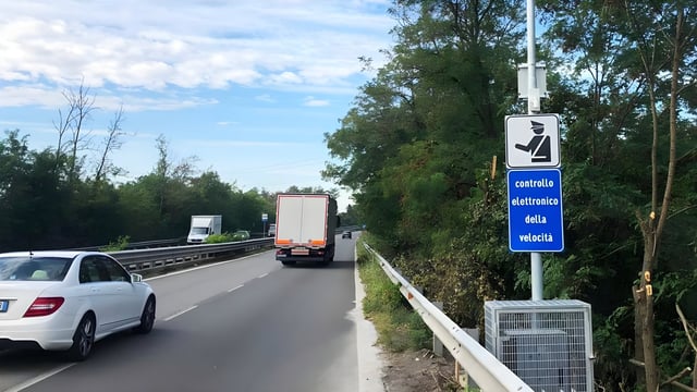 Traffic on a Milan highway passing an electronic speed control sign and roadside monitoring equipment.