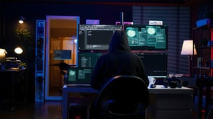 A person in a black hoodie sits at a desk facing multiple glowing computer screens displaying code
