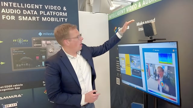 A man in a suit gestures toward a sensor and monitor at a Samiraa smart mobility data platform exhibition booth.