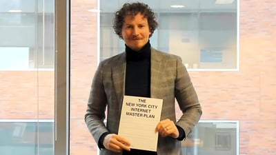 A man in a blazer holding a document titled "The New York City Internet Master Plan" in front of a window.