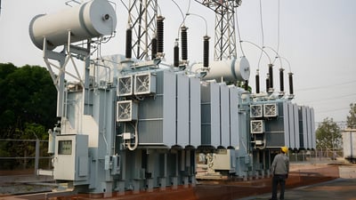 A worker in a yellow hard hat stands before large high-voltage power transformers at an electrical substation.