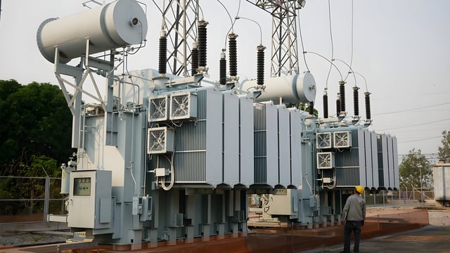 A worker in a yellow hard hat stands before large high-voltage power transformers at an electrical substation.