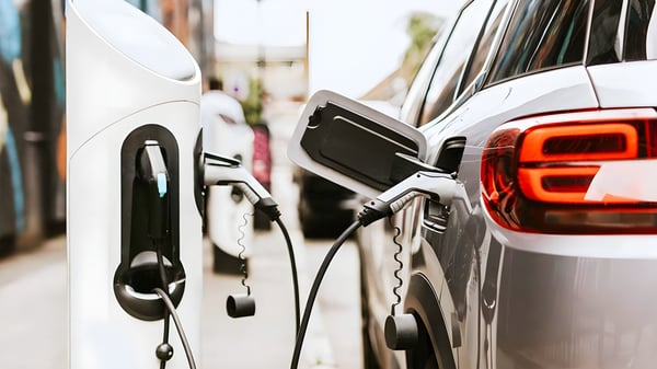 Close-up of a silver electric car plugged into a white curbside charging station on a city street.