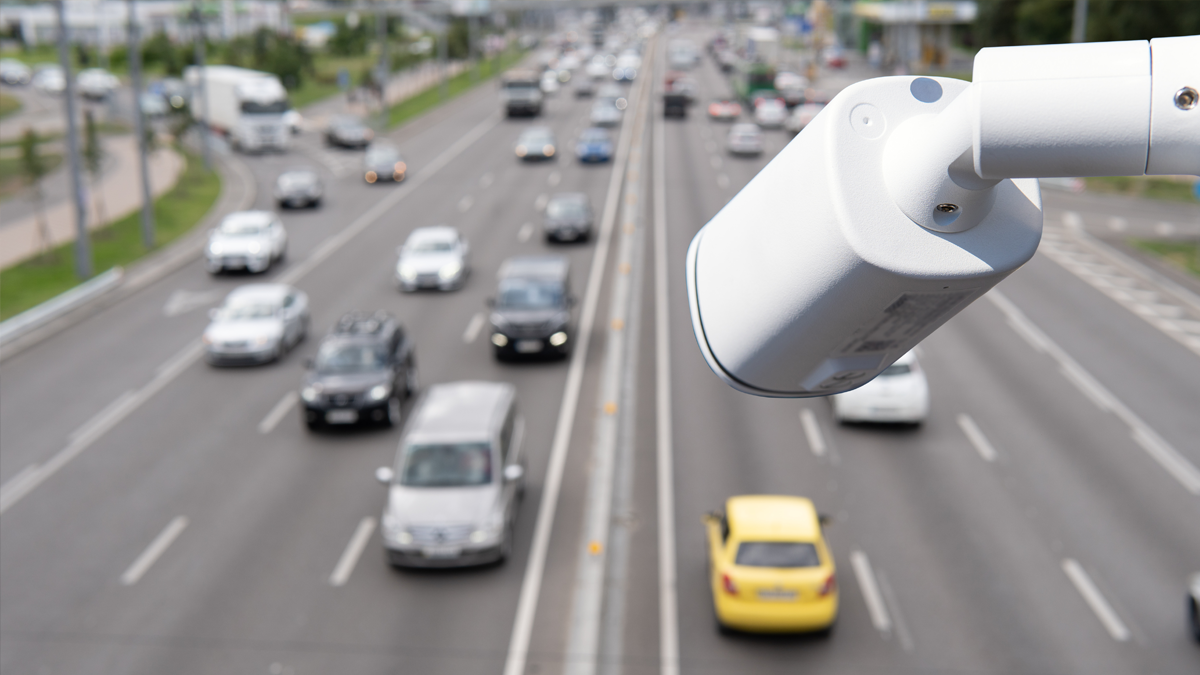 A white security camera mounted above a busy multi-lane highway overlooking blurred traffic.