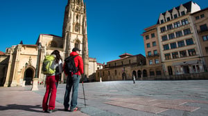 Two hikers with backpacks stand in a paved plaza facing the Cathedral of San Salvador in Oviedo, Spain.